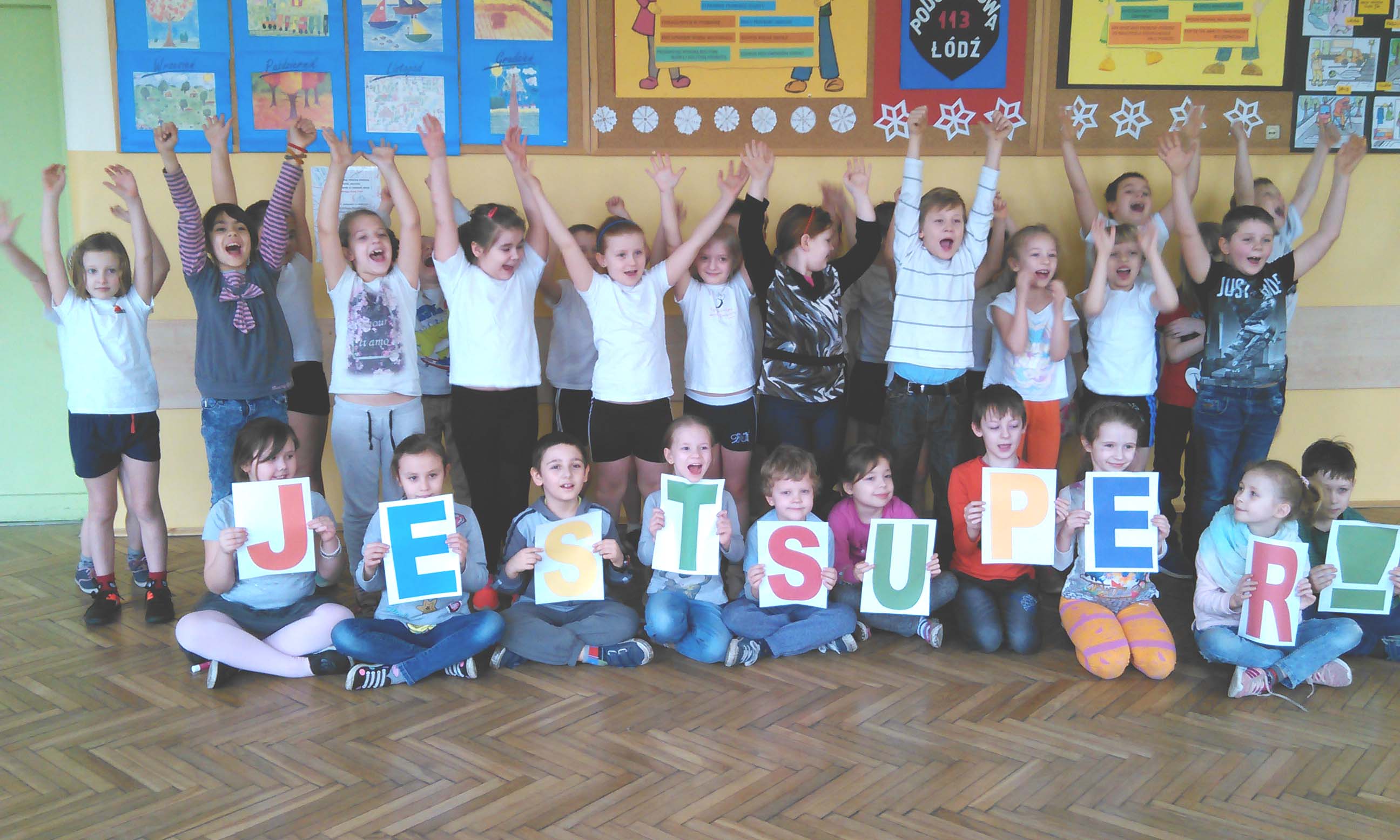 Grupa dzieci ubranych kolorowo. Stojące dzieci w tylnym rzędzie podnoszą ręce do góry. Siedzące dzieci w przednim rzędzie trzymają litery układające się w napis Jest super!