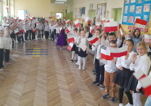 Uczniowie ubrani w stroje galowe z biało-czerwonymi chorągiewkami podczas uroczystego apelu.