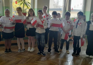 Uczniowie ubrani w stroje galowe z biało-czerwonymi chorągiewkami podczas uroczystego apelu.