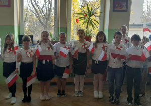 Uczniowie ubrani w stroje galowe z biało-czerwonymi chorągiewkami podczas uroczystego apelu.