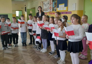 Uczniowie ubrani w stroje galowe z biało-czerwonymi chorągiewkami podczas uroczystego apelu.