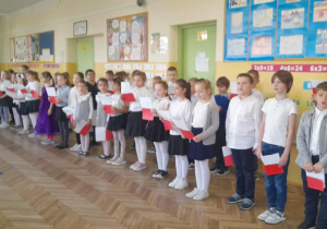 Uczniowie ubrani w stroje galowe z biało-czerwonymi chorągiewkami podczas uroczystego apelu.