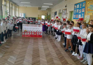 Uczniowie ubrani w stroje galowe z biało-czerwonymi chorągiewkami podczas uroczystego apelu.