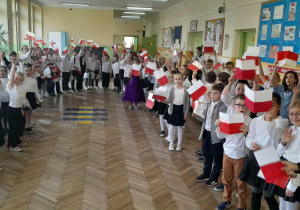 Uczniowie ubrani w stroje galowe z biało-czerwonymi chorągiewkami podczas uroczystego apelu.