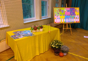 Obok tablicy stoi stół przykryty żółta tkaniną, na którym leży duży ołówek do pasowania ucznió oraz pamiątkowe dyplomy.