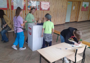 Troje uczniów wrzuca karty do głosowania do urny. Jeden uczeń zaznacza swój wybór na karcie do głosowania.
